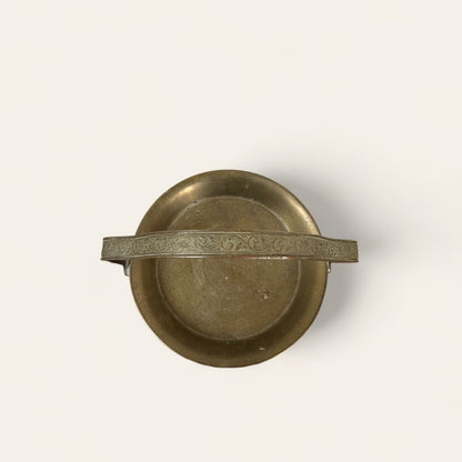 An overhead view of the Mini Brass Tray by Shoppe Maison reveals a vintage brass piece with a single horizontal handle adorned with intricate floral engravings. The tray boasts a smooth, slightly tarnished surface that highlights its aged character. Perfect for lovers of vintage goods and home decor, the background is plain and light-colored.