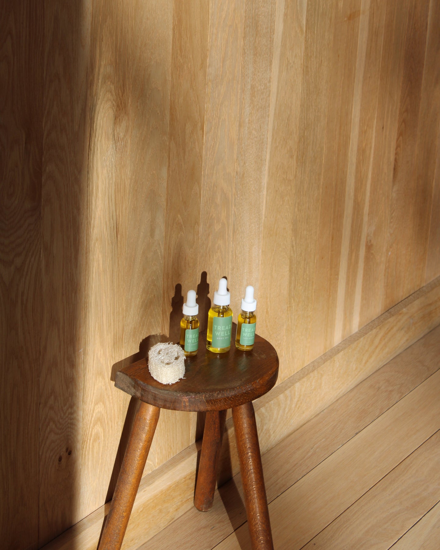 Three Mini Essentials hydrating face serums by & Tread Well and a round natural sponge rest on a rustic wooden stool in sunlight, highlighting the purity of all-natural skincare.