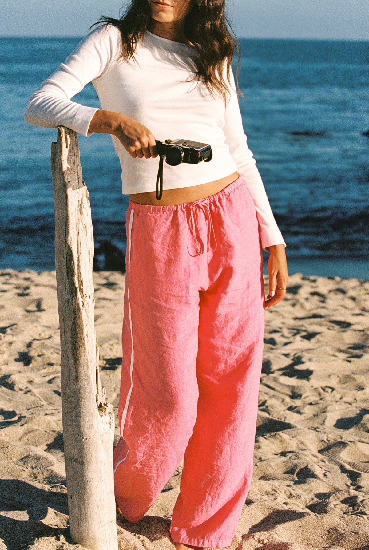 A woman wearing the LA RELAXED Linen Track Pant in pink and a white long-sleeve top leans against driftwood on a sandy beach, holding a camera with the ocean in the background.