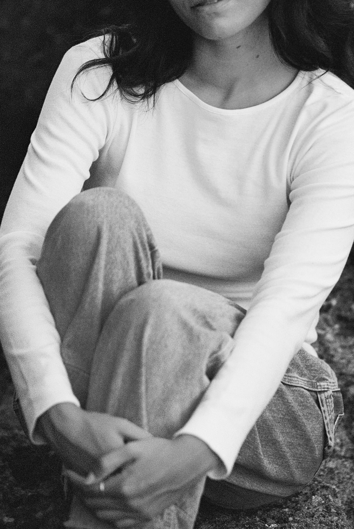 A black and white photo shows a woman sitting with her knees up, wearing the Bebe Organic Long Sleeve from LA RELAXED and jeans. Her hands are clasped in front of her legs, while part of her face is out of frame.