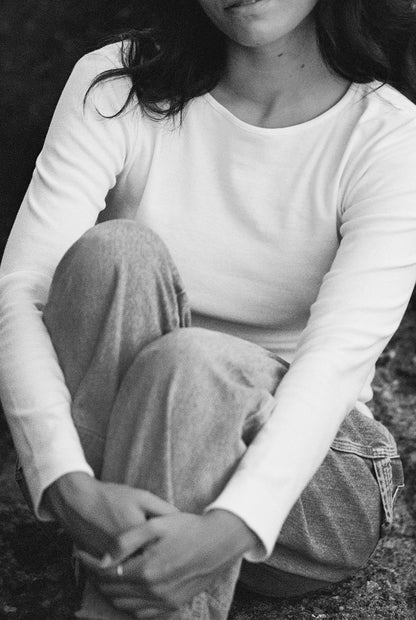 A black and white photo shows a woman sitting with her knees up, wearing the Bebe Organic Long Sleeve from LA RELAXED and jeans. Her hands are clasped in front of her legs, while part of her face is out of frame.