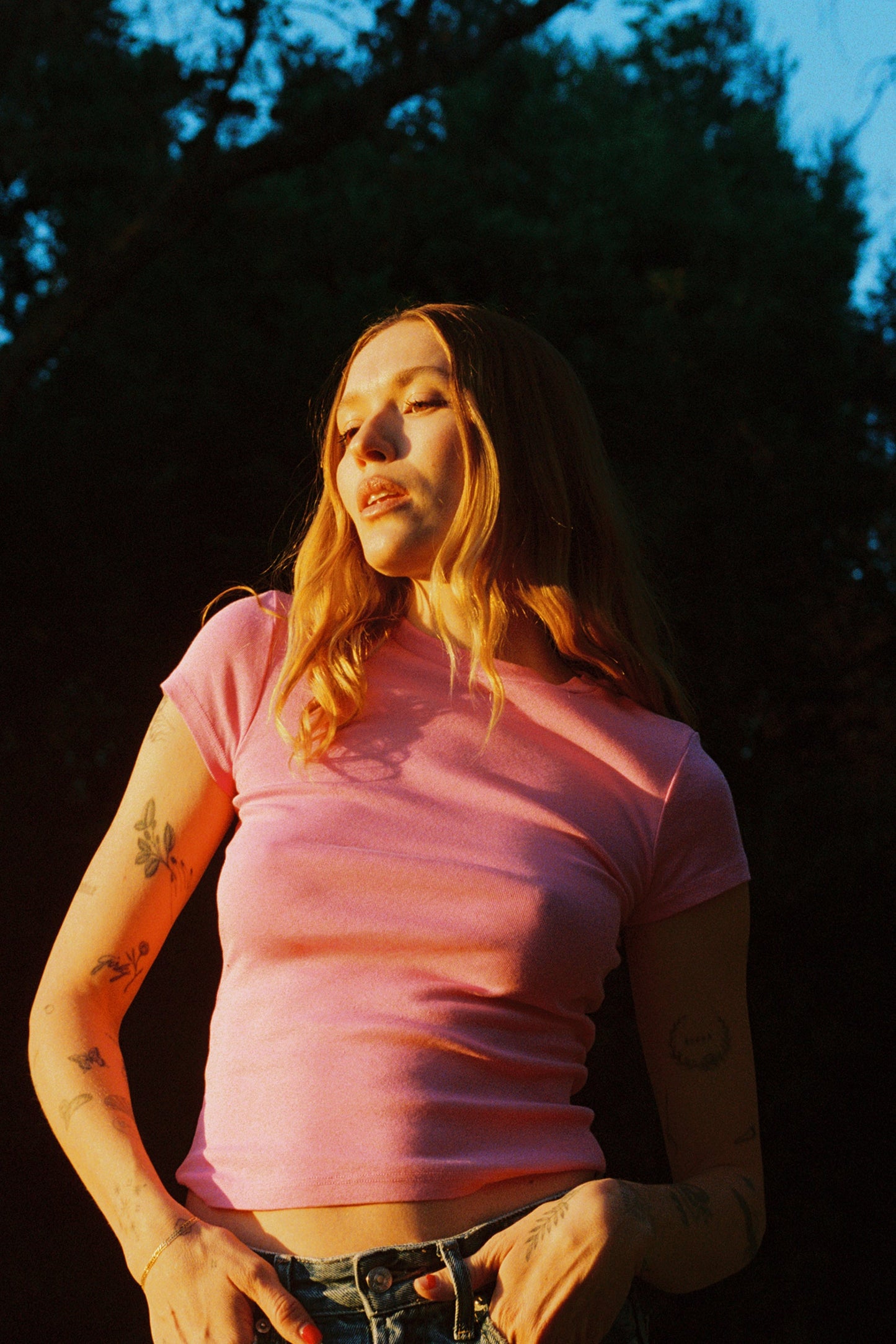 Bubblegum A person with long hair stands outdoors under the setting sun, wearing a pink LA Relaxed Bebe Organic Tee and denim jeans. Their tattooed arms are highlighted by warm lighting, with trees and a clear sky in the background.