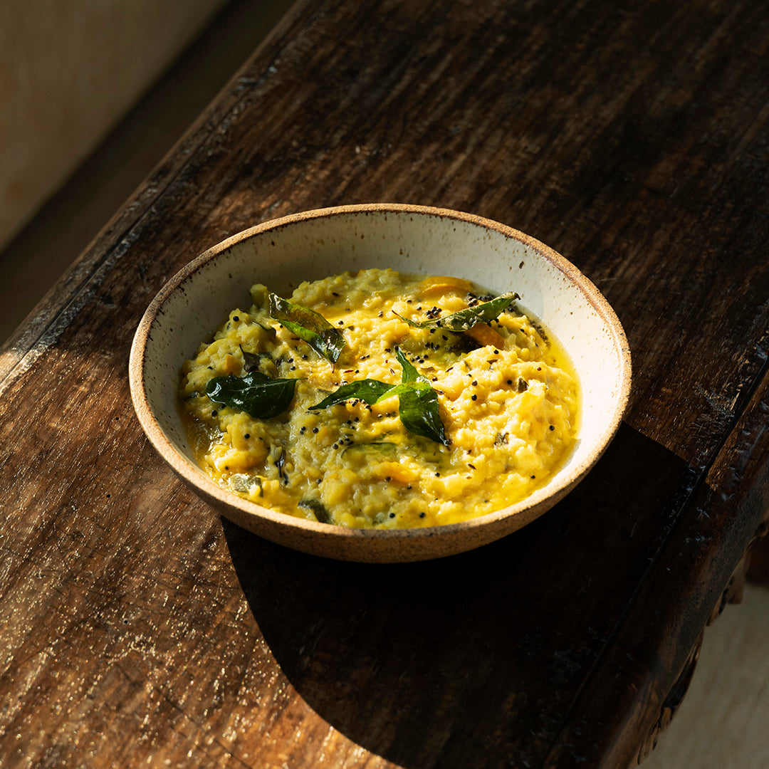 Cookma's Healing Bundle, inspired by Ayurveda, features yellow khichdi garnished with curry leaves and spices, served on a rustic wooden table bathed in natural sunlight.