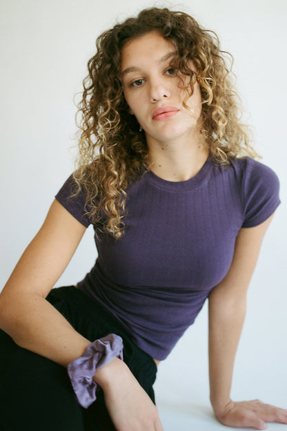 A person with curly hair wears an LA RELAXED Organic Texture Tee in purple and poses against a white background. Their wrist is adorned with a matching scrunchie, and they maintain a neutral expression while sitting slightly leaned forward.