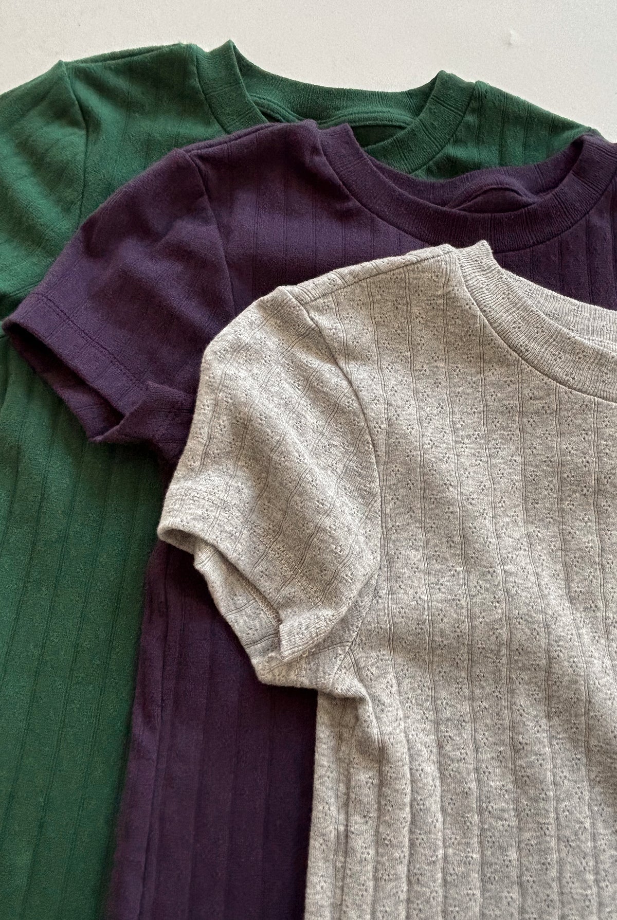Three Organic Texture Tees from LA RELAXED are arranged in overlapping layers. The short-sleeve shirts, crafted from organic cotton with a visible rib knit pattern, feature dark green, dark purple, and light gray colors from top to bottom.