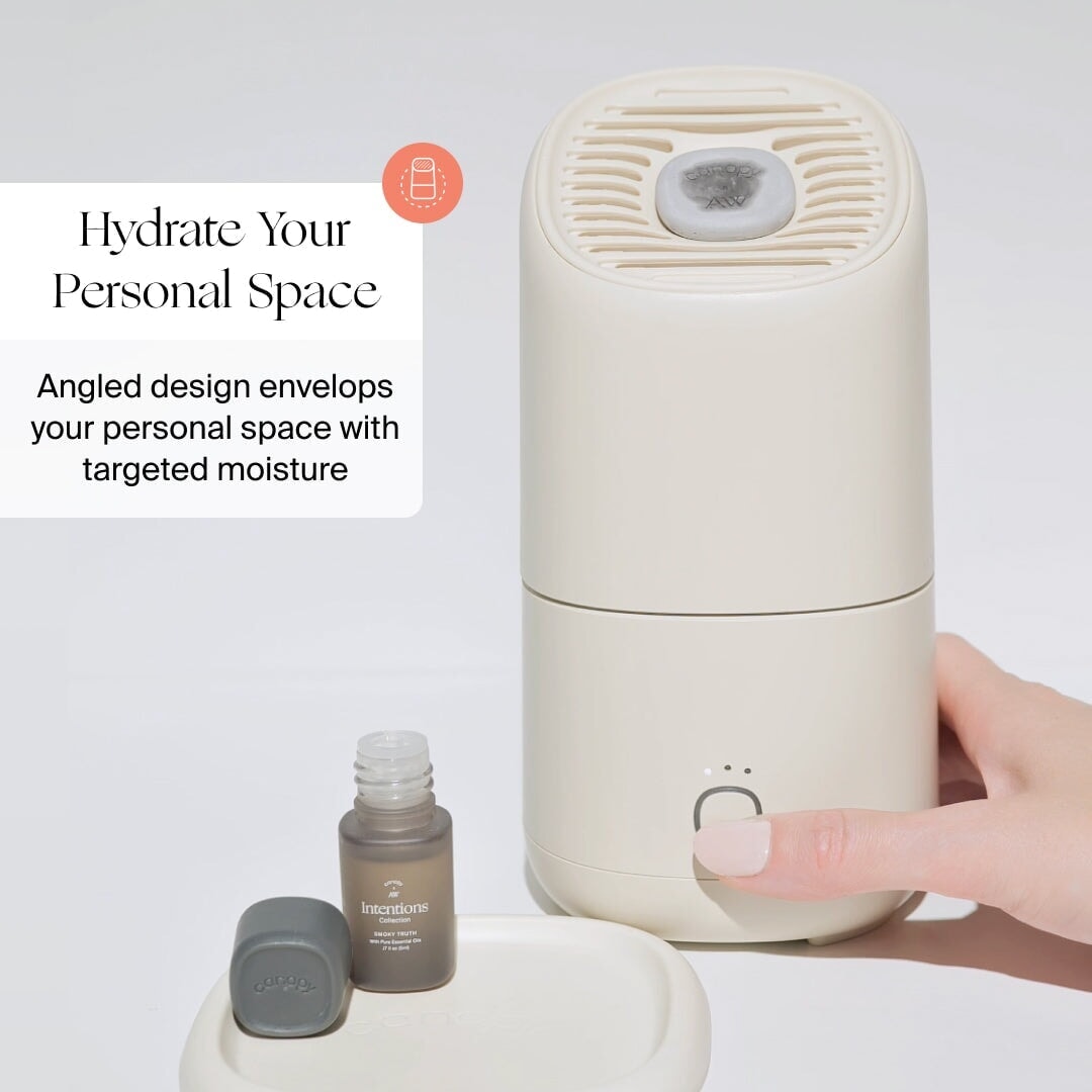 A hand presses a button on a beige Canopy Portable Humidifier beside an open essential oil bottle. Text: "Hydrate Your Personal Space. Angled design delivers targeted moisture with the Canopy Portable Humidifier.