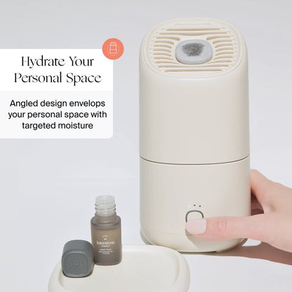 A hand presses a button on a beige Canopy Portable Humidifier beside an open essential oil bottle. Text: "Hydrate Your Personal Space. Angled design delivers targeted moisture with the Canopy Portable Humidifier.