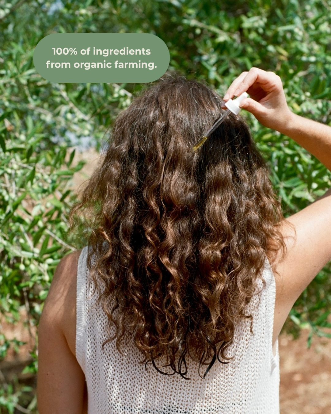 A woman with long, curly brown hair applies & Tread Well Hair Oil to her scalp with a dropper outdoors. Text reads, "100% of ingredients from organic farming." Green foliage and sunlight evoke natural repair and growth.