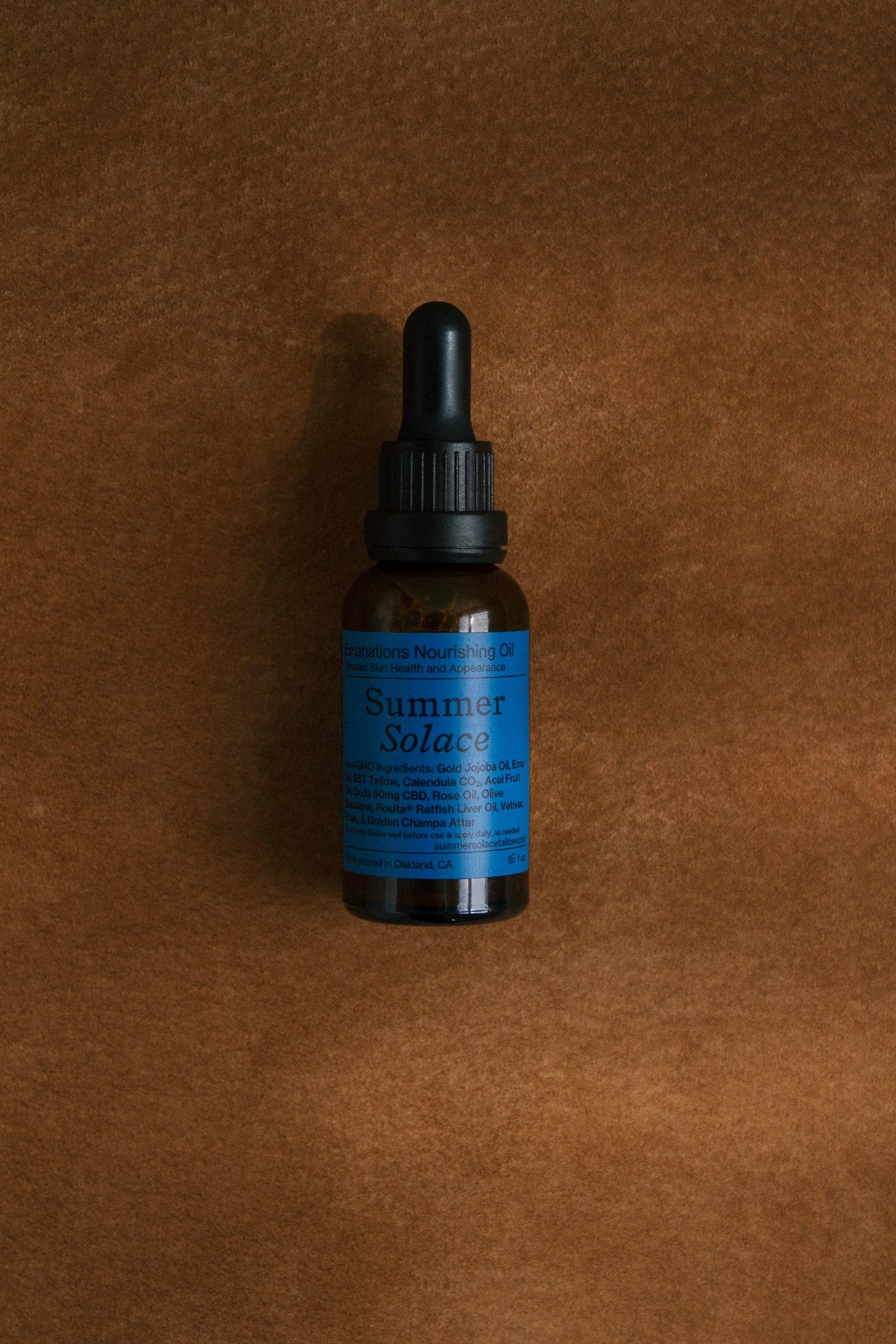 A small amber glass dropper bottle with a blue label reading "Emanations Profound Face Oil – Deeply Nourishing | Champa, Tuberose & White Ginger Lily" by Summer Solace Tallow stands upright on brown suede. Natural ingredients are listed on the label.