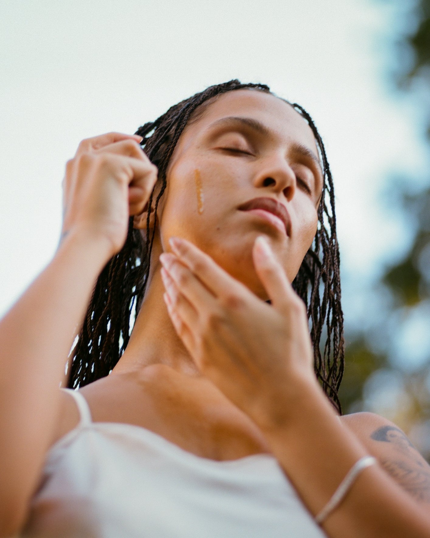 A person with braided hair wearing a white tank top is standing outdoors with eyes closed, gently applying Surya's Balancing Face Oil to their face. The background is blurry, suggesting a serene and relaxed atmosphere.