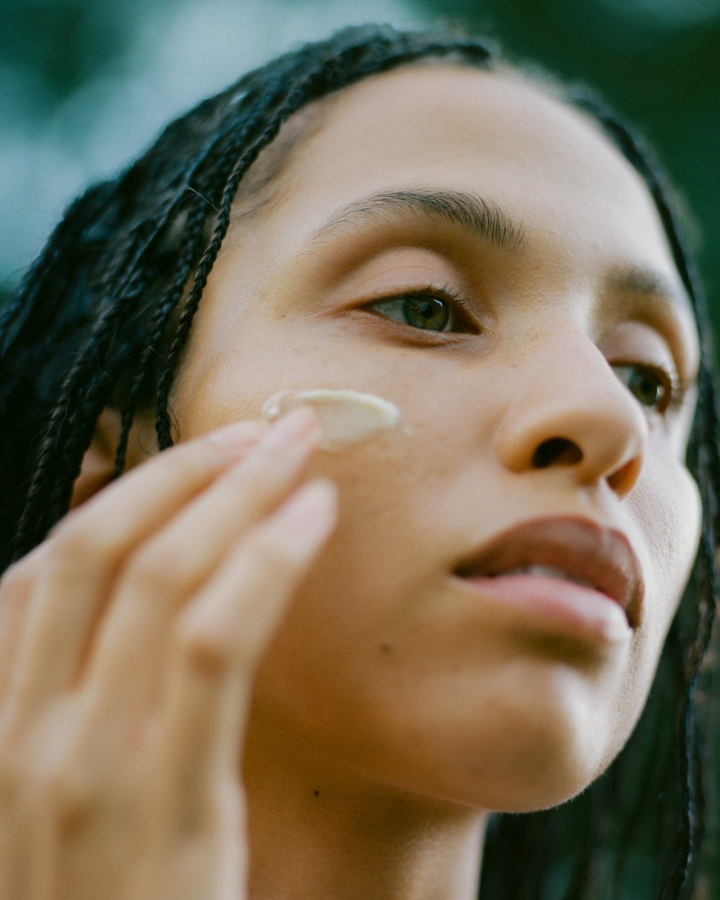 Close-up of a person with braided hair gently applying Surya's Balancing Collagen Cream to their face, displaying a peaceful expression against an out-of-focus green natural background. The image highlights skincare and self-care, enhanced by the benefits of botanical retinol.