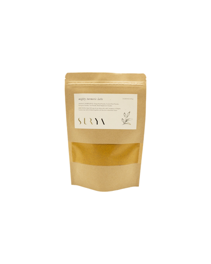 A brown kraft paper pouch labeled "Mighty Turmeric Latte" from Surya highlights its role in your wellness routine. Featuring white text, a simple plant design, and a transparent window showing the contents inside, this Ayurvedic latte mix prominently displays the brand name "Surya" on the label.