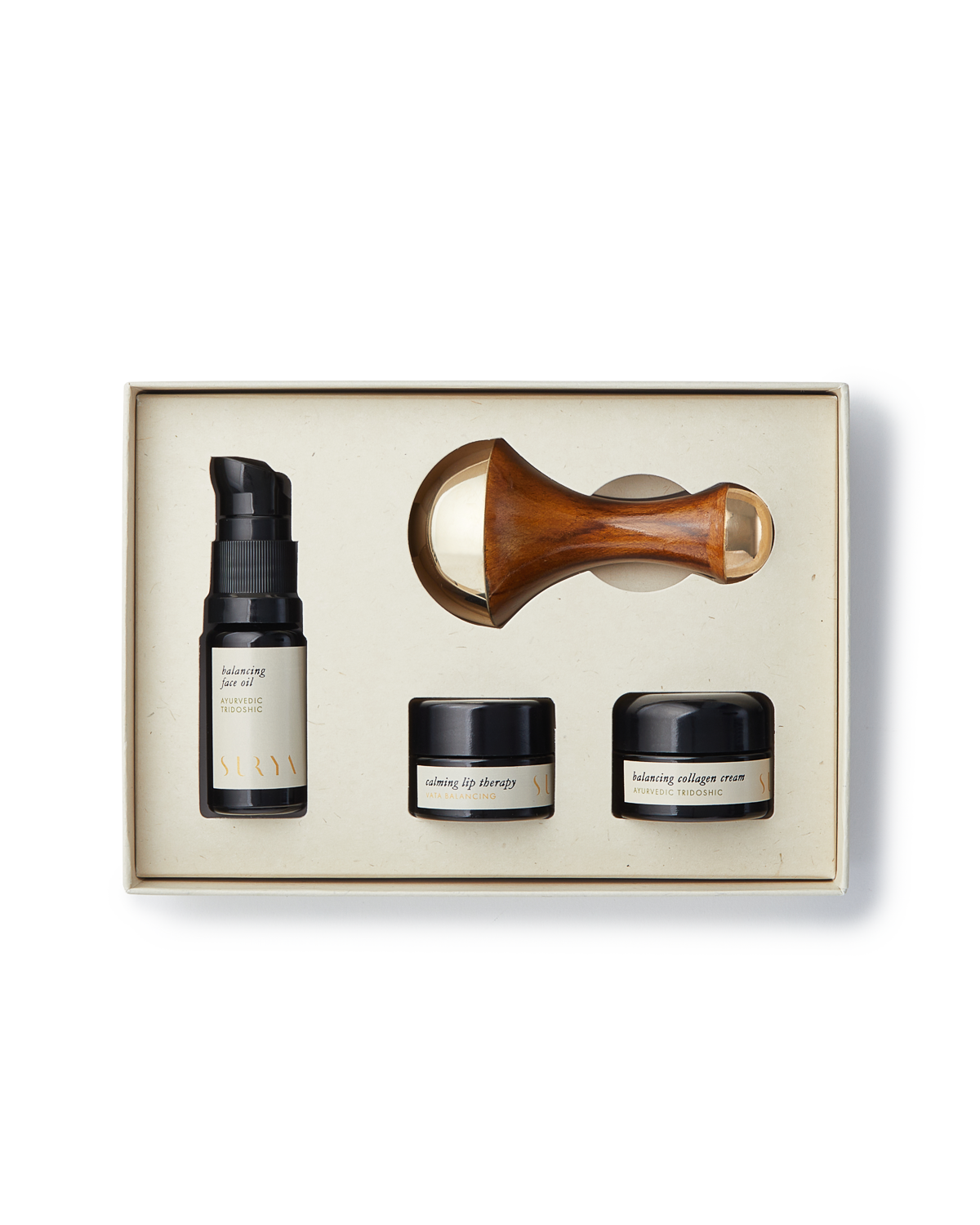 The Surya Skincare Discovery Set comes in a beige box and includes four items: a pump bottle labeled "balancing serum," two small jars labeled "lifting eye therapy" and "balancing collagen cream," and a wooden-handled Kansa Wand with a gold-colored roller head. This set provides an unparalleled Ayurvedic beauty experience.