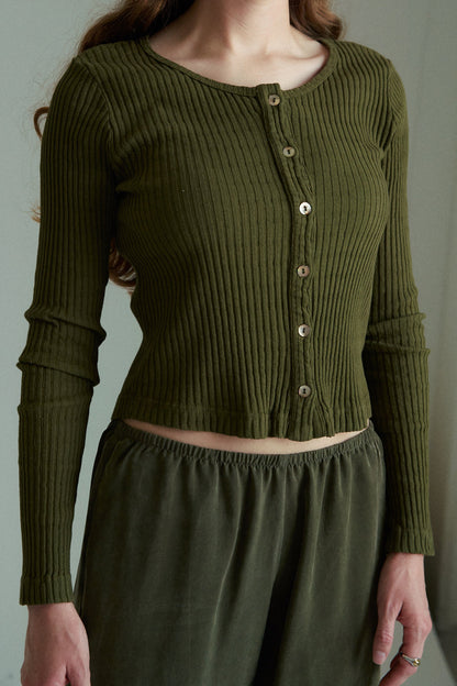 Sage- 
A woman indoors wears the LA RELAXED Pointelle Cardigan, an olive green ribbed long-sleeve top with pearlescent buttons, paired with matching loose pants. The cropped image shows her from below the shoulders without showing her face.