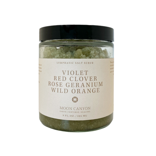 A glass jar with a black lid contains the Violet, Red Clover, Rose Geranium & Wild Orange Lymphatic Salt Scrub designed for skin health. The label reads "Moon Canyon Healing," featuring ingredients such as violet, red clover, rose geranium, and wild orange. It holds 9 fl. oz./266 ml of product, promoting effective detoxification and lymphatic drainage.