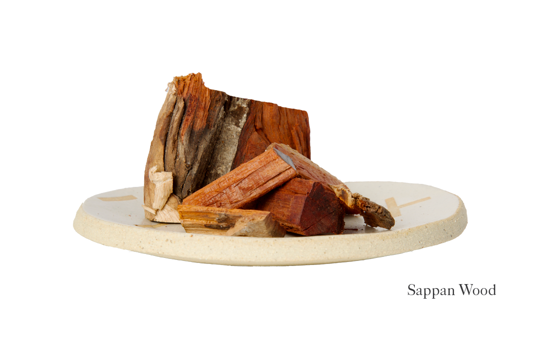 A beige ceramic plate holds several pieces of reddish-brown sappan wood, echoing the deep shade featured in the Dark Harda and Sappan Wood Ayurvedic Yoga Mat by Bennd Yoga. The varying-sized chunks are neatly stacked, while nearby, the text "Sappan Wood" highlights its earthy allure and natural beauty.