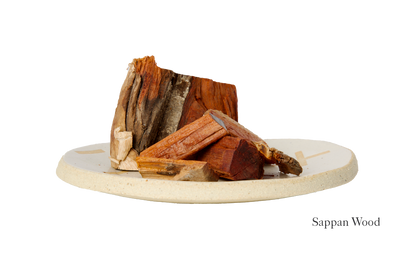 A beige ceramic plate holds several pieces of reddish-brown sappan wood, echoing the deep shade featured in the Dark Harda and Sappan Wood Ayurvedic Yoga Mat by Bennd Yoga. The varying-sized chunks are neatly stacked, while nearby, the text "Sappan Wood" highlights its earthy allure and natural beauty.