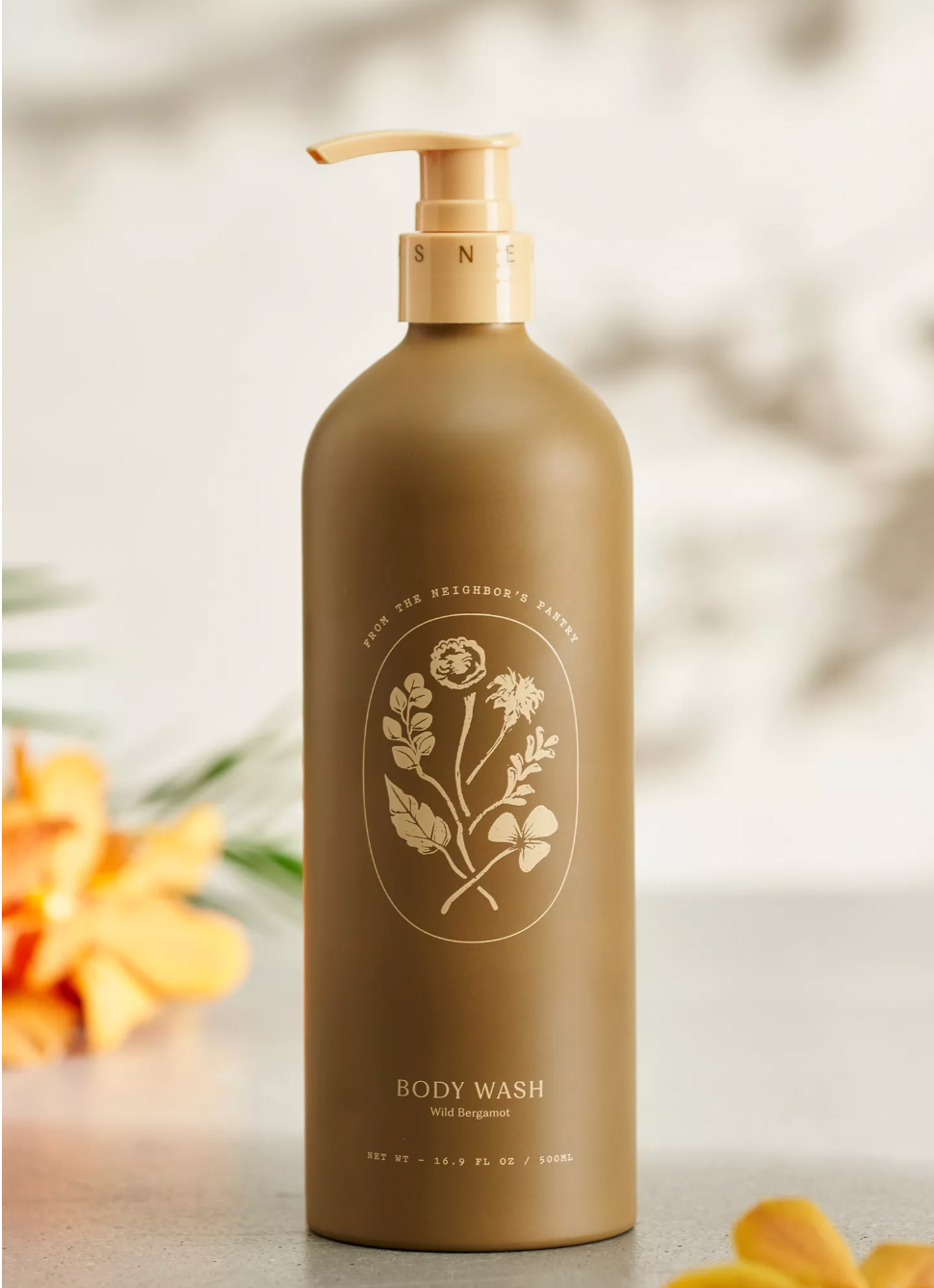 A tan bottle of My Neighbor’s Tallow Wild Bergamot Body Wash with a pump top rests on a gray surface. Its label features gold floral art and notes organic ingredients. Blurred orange and yellow flowers appear in the background.