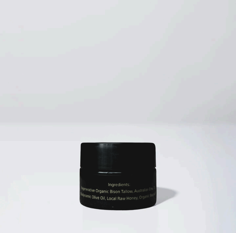 A small black jar of Wild Mamas’ Wildly Nourishing Bison Tallow Balm - Raw Honey sits on a plain white surface against a light gray background. The minimal, well-lit scene highlights the purity of this tallow skincare product.