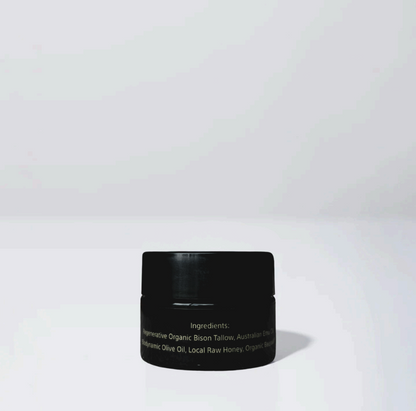 A small black jar of Wild Mamas’ Wildly Nourishing Bison Tallow Balm - Raw Honey sits on a plain white surface against a light gray background. The minimal, well-lit scene highlights the purity of this tallow skincare product.