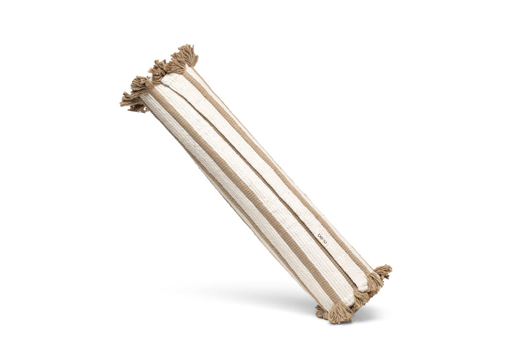 A beige and white striped Harda Ayurvedic Yoga Mat by Bennd Yoga, crafted from organic cotton, is positioned at an angle. It features fringed tassels at both ends and a textured fabric surface. The mat is displayed against a plain white backdrop, exuding a sense of natural elegance.