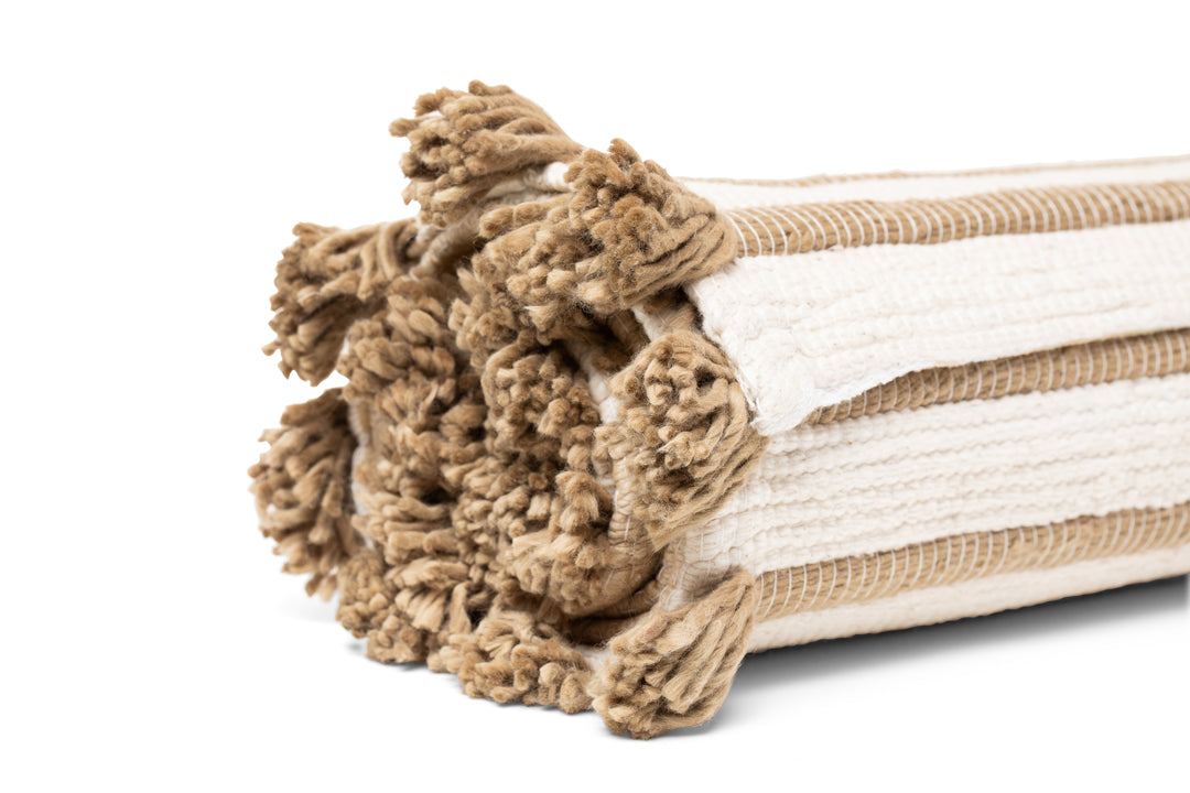 A rolled-up, woven beige and white Harda Ayurvedic Yoga Mat by Bennd Yoga features fringed tassels at the ends and a textured pattern. Crafted from organic cotton, it embodies a natural elegance. The mat is positioned on its side against a white background, exuding simplicity and charm.