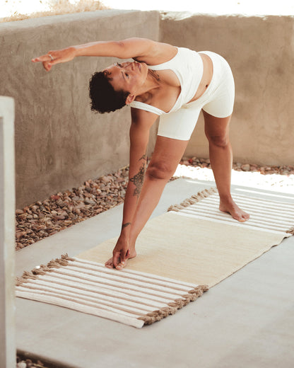Dressed in a white athletic outfit, a person gracefully performs a side stretch on the Harda Ayurvedic Yoga Mat by Bennd Yoga. Outdoors, amidst a concrete wall and gravel, they extend one arm over their head. With an arm tattoo visible, they embrace the grounded and serene feeling provided by the organic cotton surface.