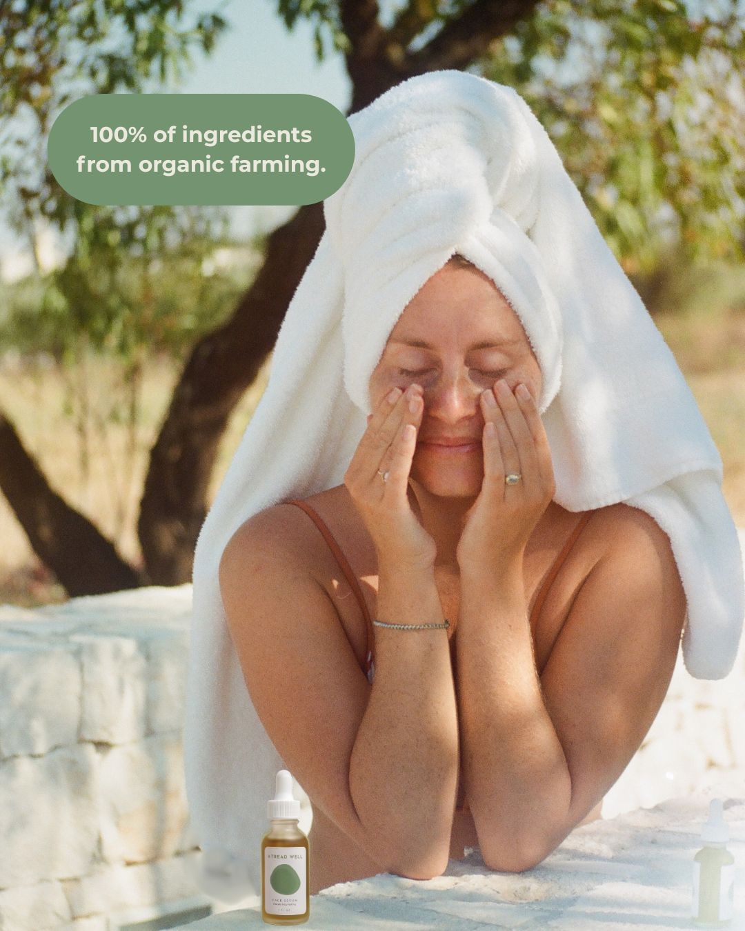 A woman with a towel on her head massages her face outdoors using & Tread Well Face Serum. A bottle is in the foreground, and a text bubble reads, "100% of ingredients from organic farming." Sunlight filters through the trees.