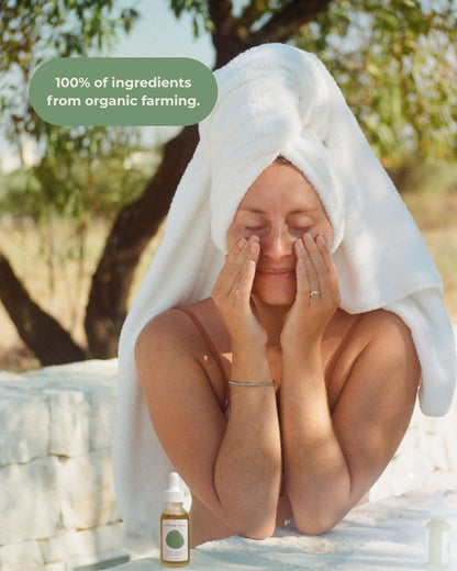 A woman with a towel on her head massages her face outdoors using & Tread Well Face Serum. A bottle is in the foreground, and a text bubble reads, "100% of ingredients from organic farming." Sunlight filters through the trees.