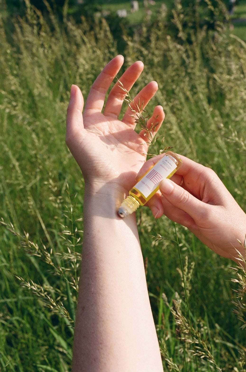 A person applies & Tread Well's Natural Fragrance - Nectar from a small glass roller bottle onto their forearm in a grassy field, enjoying its spicy cardamom scent as sunlight and green plants fill the background.