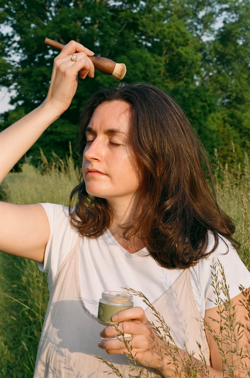 A woman with shoulder-length brown hair applies & Tread Well Dry Shampoo to her forehead with a brush while standing in a grassy field, eyes closed, wearing a white shirt and light dress. Trees fill the background.
