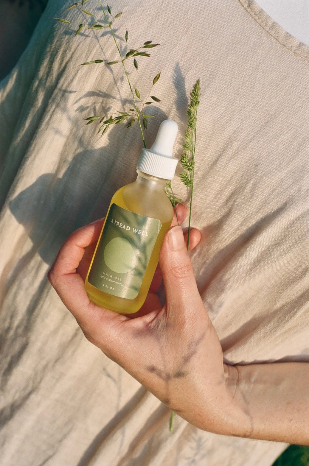 A hand holds a small glass dropper bottle of "& Tread Well" Hair Oil, perfect for repairing damage, against a beige fabric background with hints of green grass.