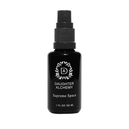 A black spray bottle labeled "Daughter Alchemy" with the text "Supreme Space® Botanical Protective Spray" and "1 FL OZ / 30 ML" near the bottom. The label, adorned with a stylized logo encircled by a wreath design, indicates the inclusion of organic white sage. The bottle has a black cap and a spray nozzle.