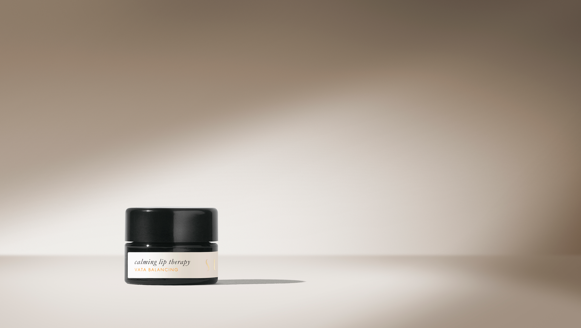 A small black jar labeled "Surya Calming Lip Therapy" sits on a beige surface. The background displays a gradient of soft beige tones, highlighted by a beam of light that casts a shadow behind the jar. This moisturizing blend features organic ingredients and boasts a creamy formula for ultimate care.