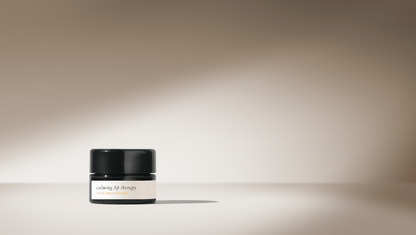 A small black jar labeled "Surya Calming Lip Therapy" sits on a beige surface. The background displays a gradient of soft beige tones, highlighted by a beam of light that casts a shadow behind the jar. This moisturizing blend features organic ingredients and boasts a creamy formula for ultimate care.