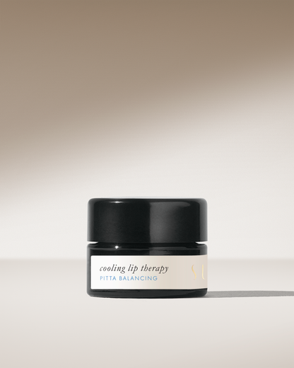 A minimalist image features a small, black jar with a beige label. The label reads "Cooling Lip Therapy" and "Pitta Balancing" in light text. Made by Surya from organic ingredients, this natural blend of lip balm casts a shadow against a beige background, creating a clean and elegant presentation.