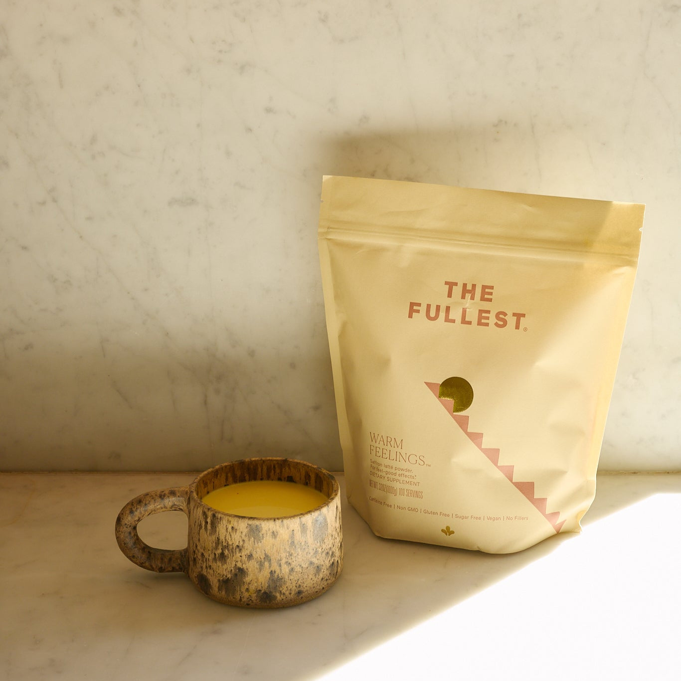 A rustic ceramic mug filled with a yellow Saffron Latte 100-Day Supply by THE FULLEST sits on a marble surface beside its beige bag, while sunlight casts soft shadows behind them.