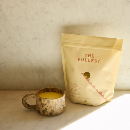 A rustic ceramic mug filled with a yellow Saffron Latte 100-Day Supply by THE FULLEST sits on a marble surface beside its beige bag, while sunlight casts soft shadows behind them.