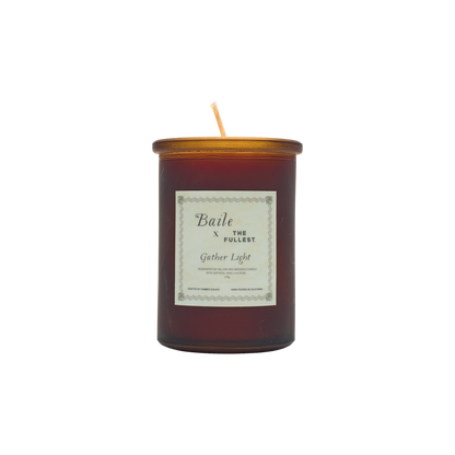 A brown glass jar candle called "Gather Light - Saffron Beeswax Tallow Candle" by Baile features a gold lid and white label. The unlit, centered wick is non-toxic, and the product is shown against a plain white background.