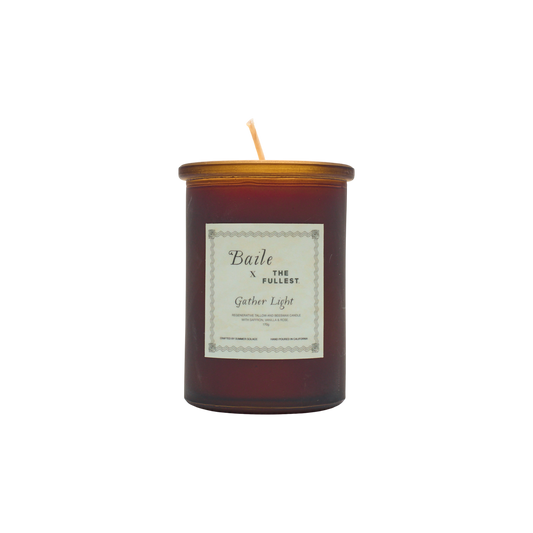 A brown glass jar candle called "Gather Light - Saffron Beeswax Tallow Candle" by Baile features a gold lid and white label. The unlit, centered wick is non-toxic, and the product is shown against a plain white background.