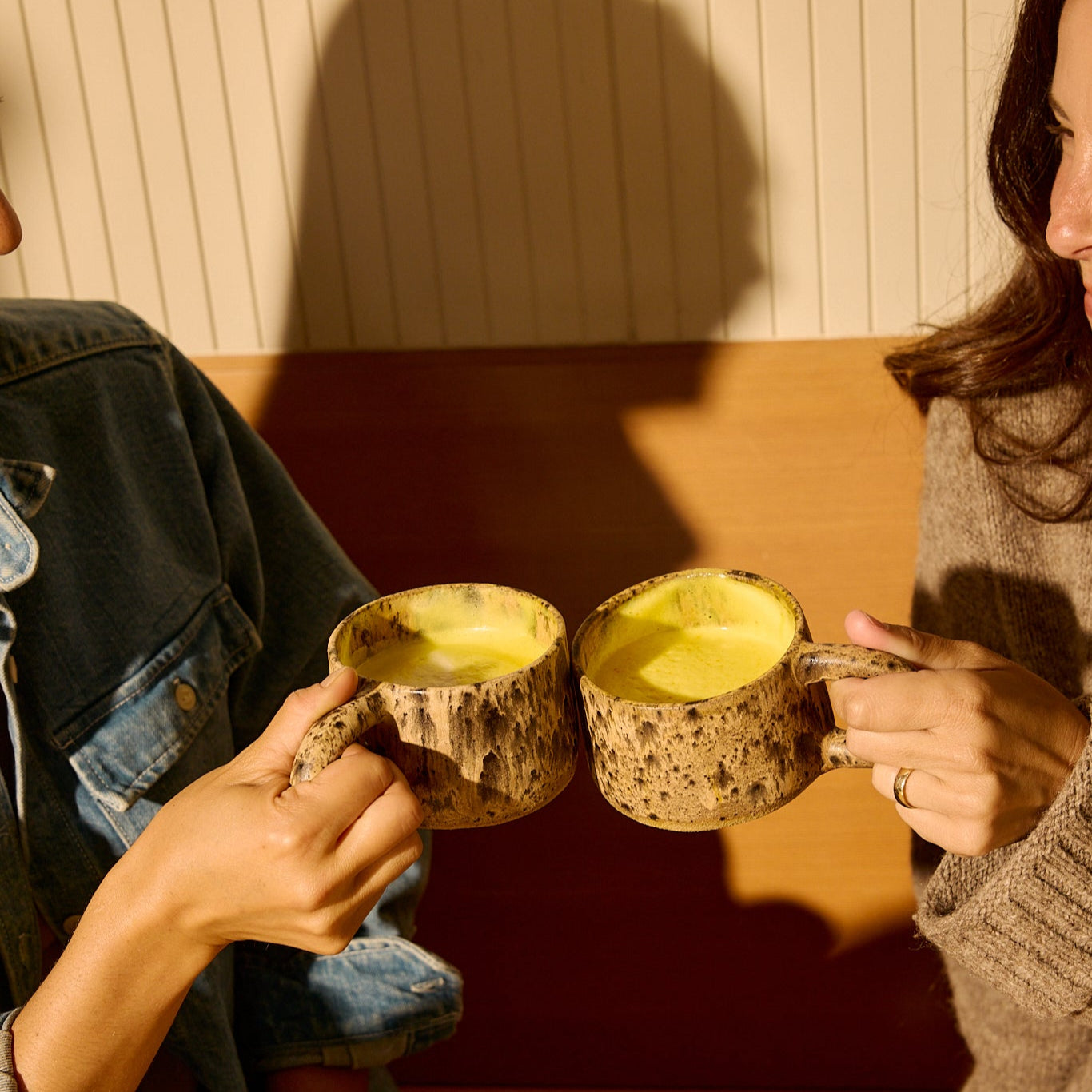 Two people clink rustic mugs filled with THE FULLEST’s Saffron Latte 100-Day Supply, a caffeine-free yellow drink known as a mood booster, while sitting on a wooden bench in a cozy, warmly lit setting.