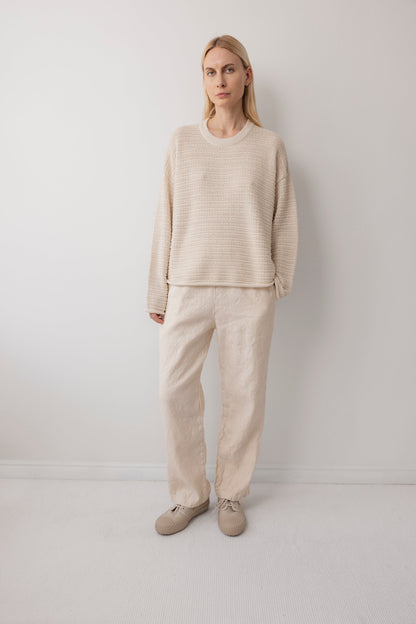 A woman with straight blonde hair and a neutral expression stands against a white wall, wearing Wol Hide's Thermal Sweater in Natural, paired with light beige loose pants and beige slippers.