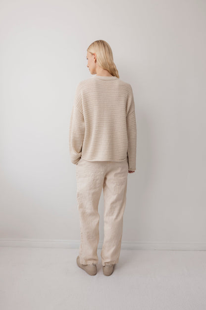 A woman with long blonde hair stands facing a white wall, wearing the Wol Hide Thermal Sweater in Natural, paired with loose cream-colored pants and light shoes. The minimalist setting features a white floor and wall.