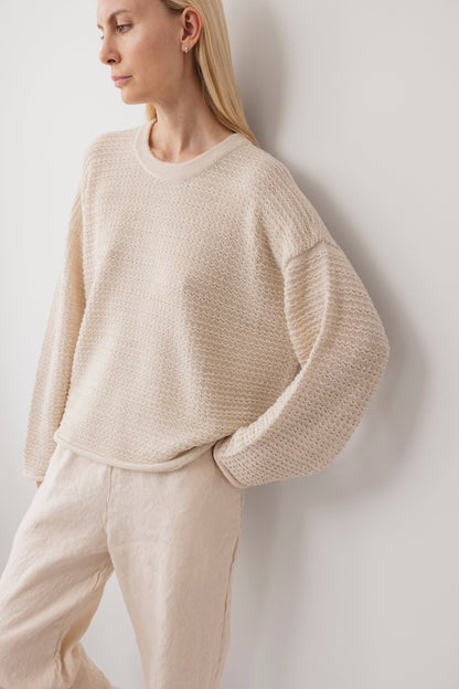 A person wears the Wol Hide Thermal Sweater in Natural and light beige pants, standing against a white wall with hands in pockets. The look is relaxed and minimalist.