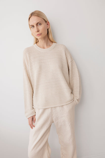 A woman with long blonde hair, wearing the Wol Hide Thermal Sweater in Natural and loose cream-colored pants, stands calmly against a plain white wall, looking at the camera.