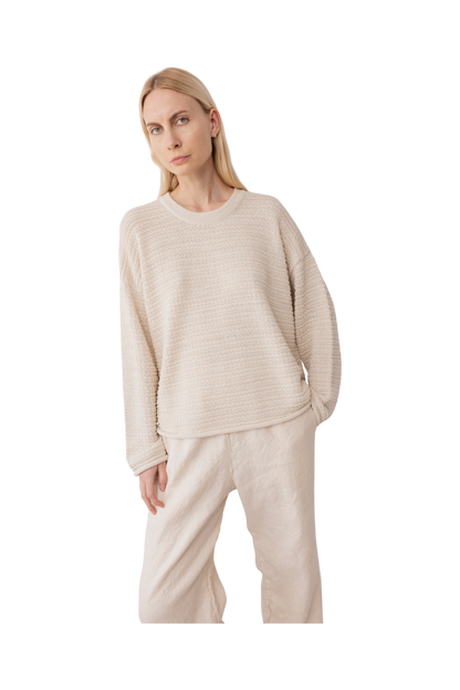 A woman with long blonde hair wears the Wol Hide Thermal Sweater in Natural and wide-leg beige pants, standing against a plain white background and capturing the spirit of slow fashion.