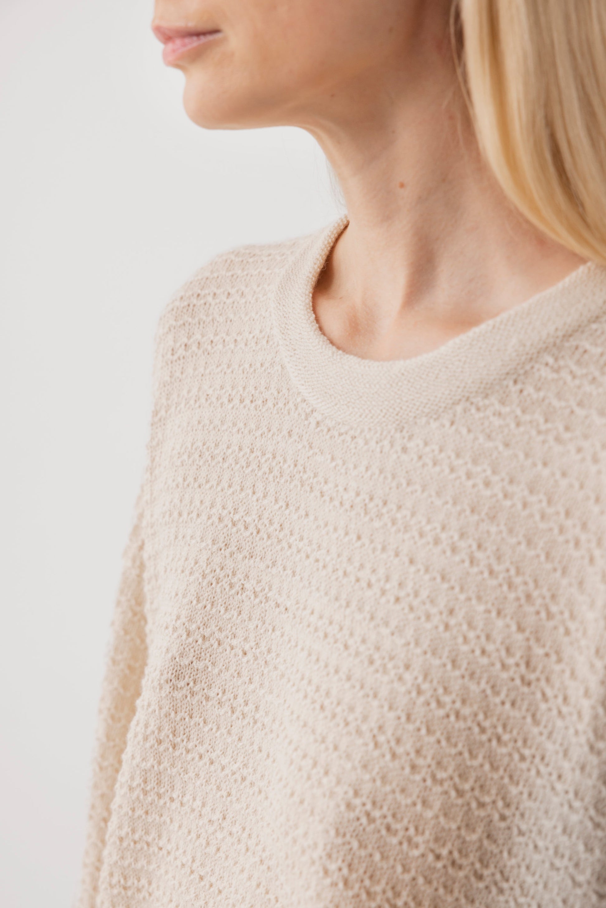 A close-up of a person wearing the Wol Hide Thermal Sweater : Natural, a lightweight, textured beige pullover made from regenerative yarn. The neckline and shoulder are shown, with long blonde hair against a plain white backdrop.