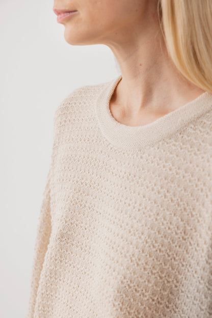 A close-up of a person wearing the Wol Hide Thermal Sweater : Natural, a lightweight, textured beige pullover made from regenerative yarn. The neckline and shoulder are shown, with long blonde hair against a plain white backdrop.
