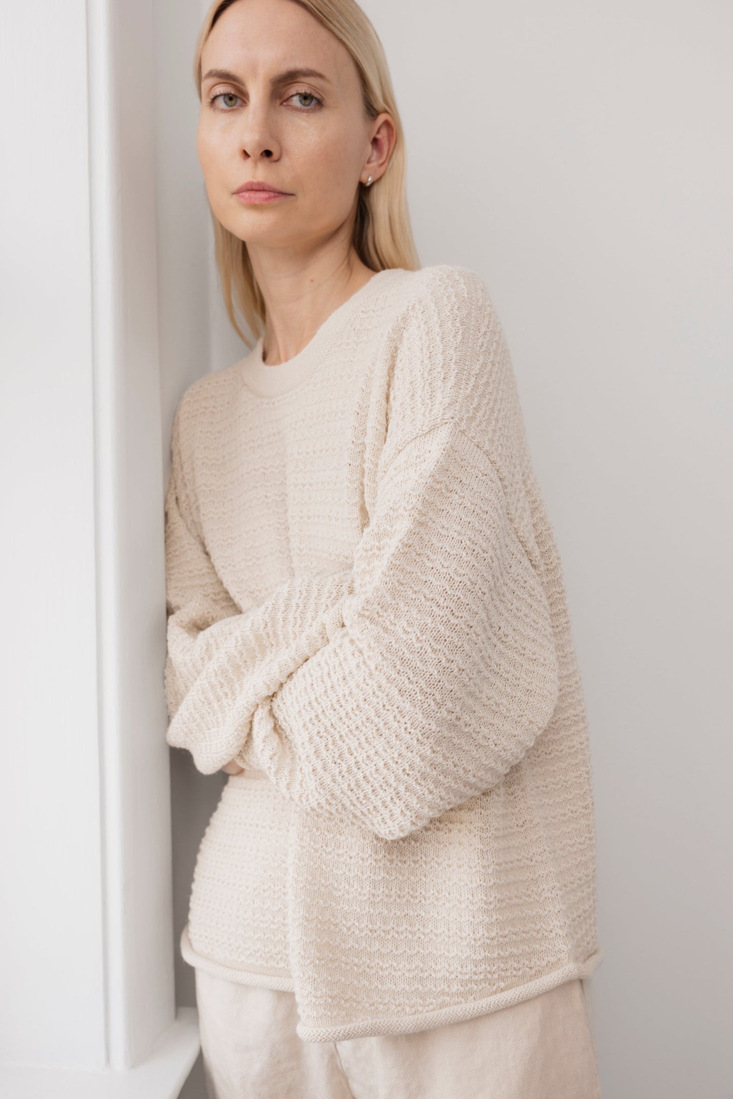 A woman with straight blonde hair stands by a white wall, calmly looking at the camera with arms loosely crossed, wearing the Wol Hide Thermal Sweater in Natural.