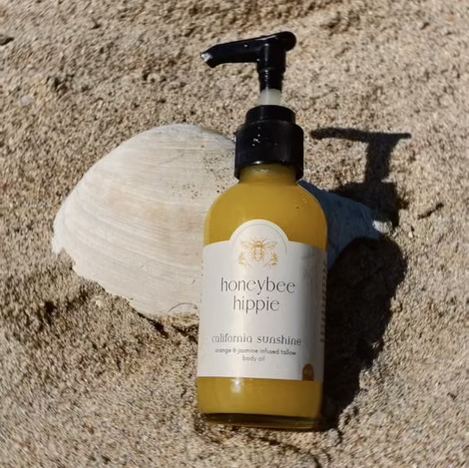 A bottle of Honeybee Hippie's "California Sunshine - Tallow Oil" with a pump rests on sandy ground, casting a shadow. Infused with organic orange peel, its non-comedogenic formula keeps skin smooth and clear. A large seashell in the background adds coastal charm.